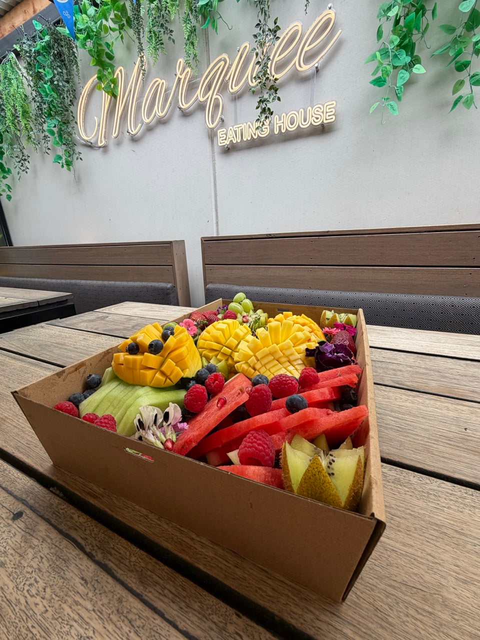 A catering box filled with assorted fresh fruit, including sliced mango, watermelon, kiwi, grapes, strawberries, blueberries, and raspberries, arranged on a weathered wooden surface.